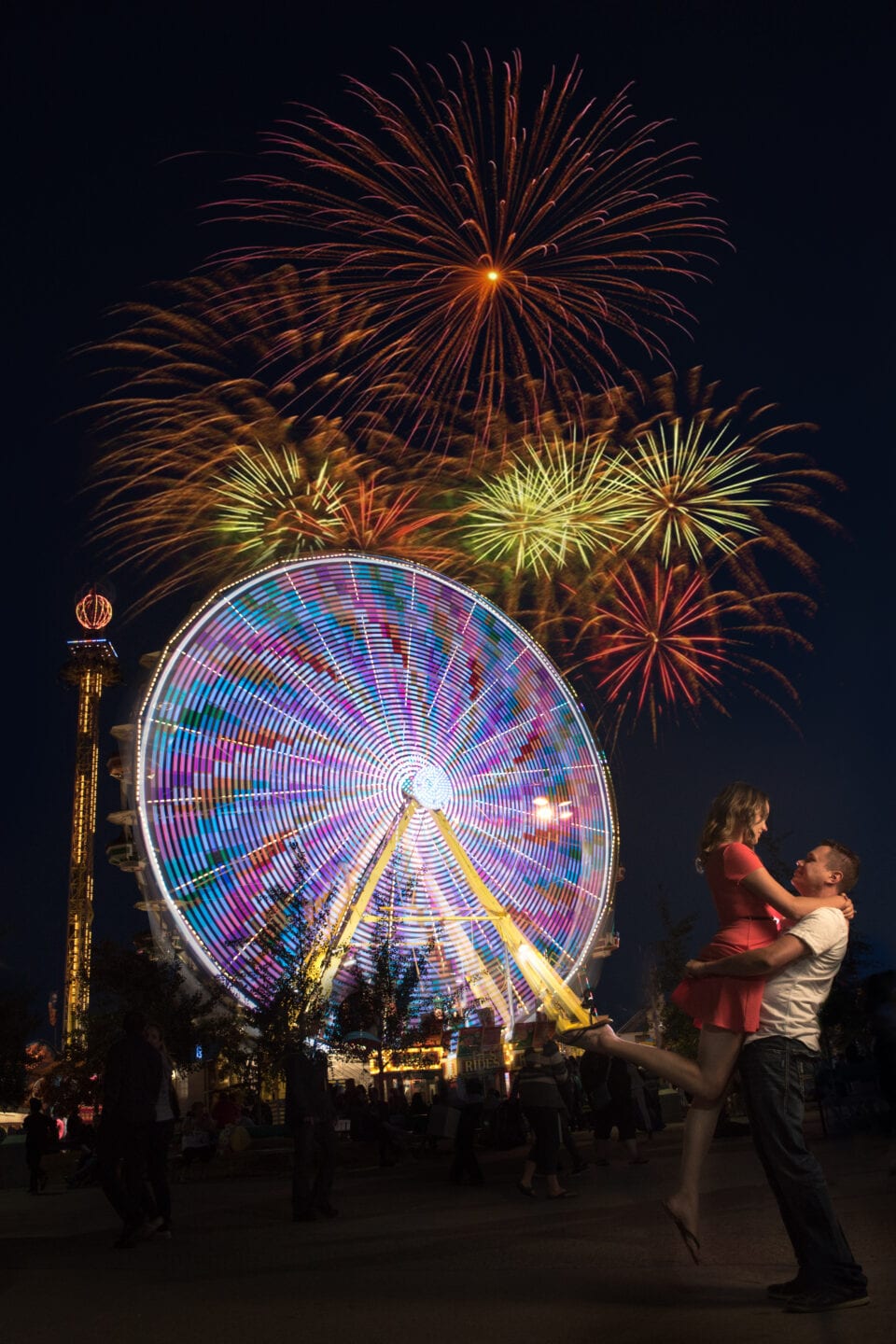 fireworks displaying on top of a ferris wheel while a couple embrace one another